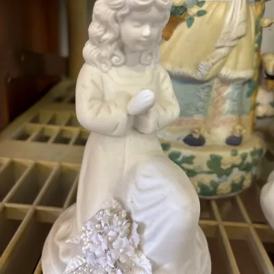 White bisque porcelain figurine of a praying girl adorned with faux pearls and fabric flowers at her base.