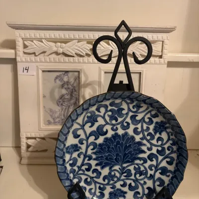 Blue and white porcelain plate with intricate floral and scroll motifs, displayed on a black wrought iron easel in front of a carved...