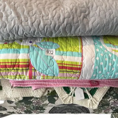 Stack of folded bedding and textiles including quilted throws, a colorful patterned quilt, a pink blanket, and a crocheted Afghan,...