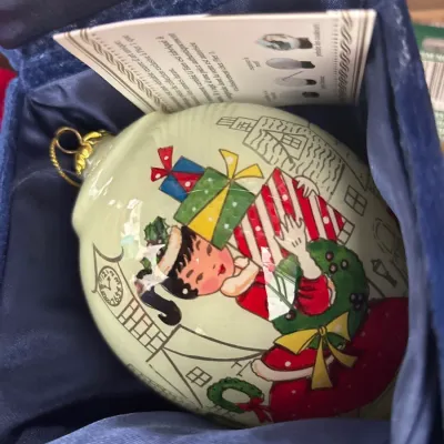 Hand-painted Li Bien reverse-painted glass Christmas ornament depicting a holiday shopper, presented in its original navy blue velvet...