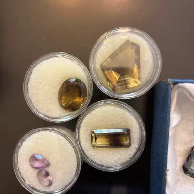 Assortment of faceted loose gemstones, including citrine, smoky quartz, and light amethyst cuts, displayed in round padded acrylic jars.