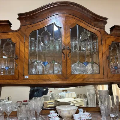 Antique wooden hutch featuring leaded glass doors with oval and stained glass accents, set over a mirrored back console displaying...