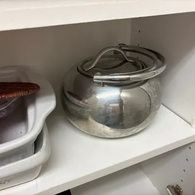 Vintage stainless steel tea kettle sitting on a cabinet shelf next to white ceramic baking dishes.