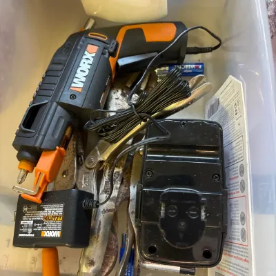 Assorted tools and accessories inside a clear storage bin, including a Worx cordless screwdriver, battery charger, and locking pliers.