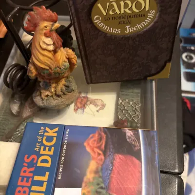 A small painted rooster figurine displayed alongside books, including Weber's Art of the Grill Deck.