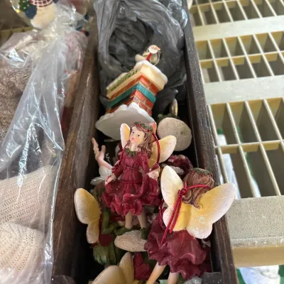 Collection of Christmas decorations inside a dark wooden trough, featuring fairy ornaments with wings and red dresses, along with a snow...