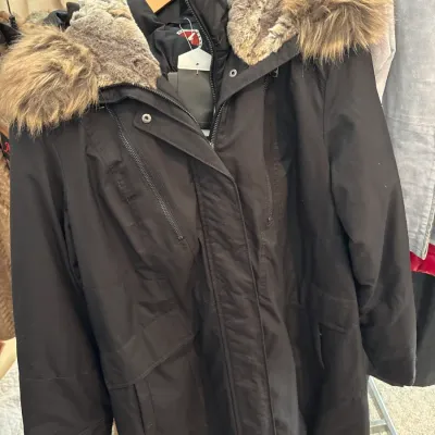 Black winter parka coat featuring a faux fur-trimmed hood and multiple zippered pockets, hanging on a clothing rack.