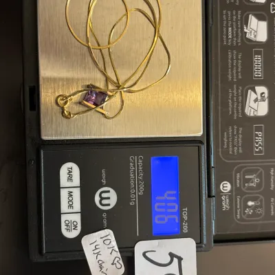 Gold necklace featuring a 14K gold chain and a 10K gold pendant set with a square-cut purple stone, shown on a digital scale weighing...