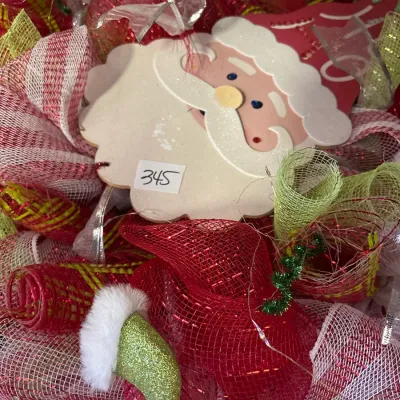 Festive Christmas wreath made of red, white, and green mesh ribbon, featuring a central wooden Santa Claus face embellishment.