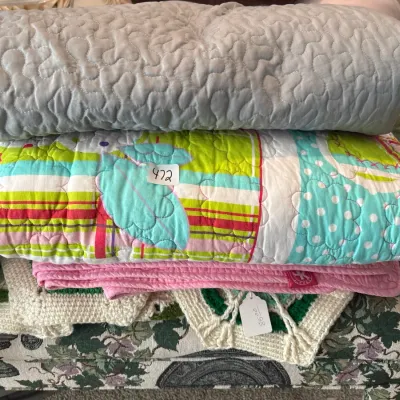 Stack of folded quilts and blankets, including a gray quilted throw, a colorful floral and striped pattern quilt, a pink blanket, and a...
