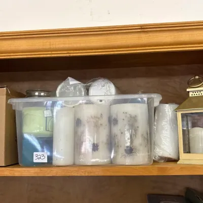 A collection of various pillar candles, jar candles, and a small brass-toned lantern arranged inside a clear plastic bin on a wooden shelf.