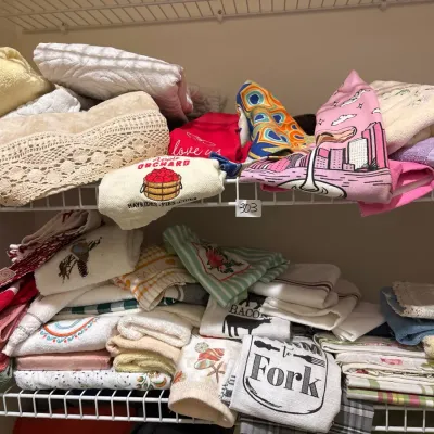 Assorted collection of folded kitchen hand towels, dish towels, and embroidered linens stacked on wire closet shelves.