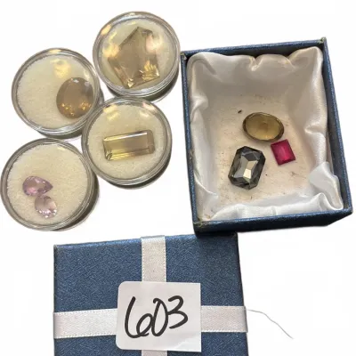 Collection of assorted faceted gemstones in clear display cases and a blue presentation box.