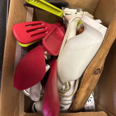 Cardboard box filled with assorted kitchen utensils, including a white handheld immersion blender and various colorful silicone spatulas.