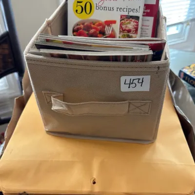 Beige fabric storage bin filled with recipe booklets and magazines, resting on top of a stack of Office Depot clasp envelopes.