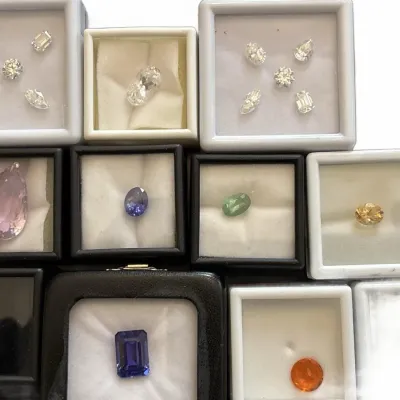 Collection of loose gemstones in individual display cases, including clear stones in various cuts, a large pear-cut pink gemstone, and...
