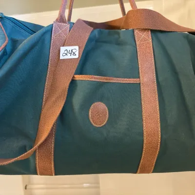 Vintage dark green canvas duffel bag featuring brown leather trim, dual top handles, an adjustable shoulder strap, and an embossed...