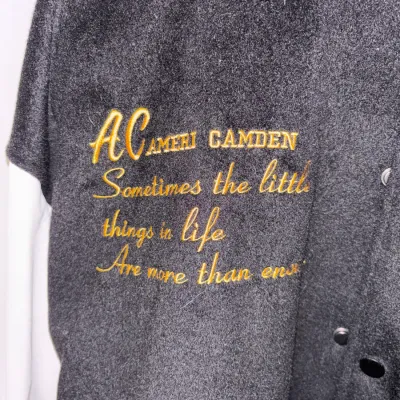 Detail of custom embroidered text in gold thread on a black felt varsity jacket.