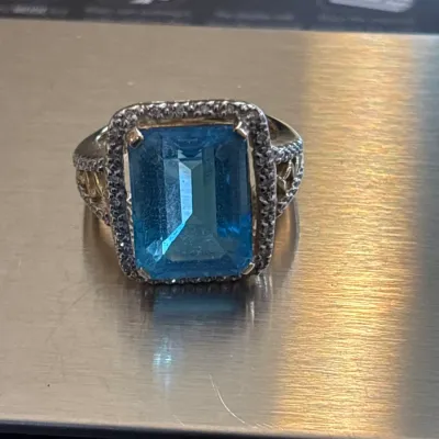 Large emerald-cut blue topaz cocktail ring set in sterling silver with a pave halo surround and ornate side filigree detail.