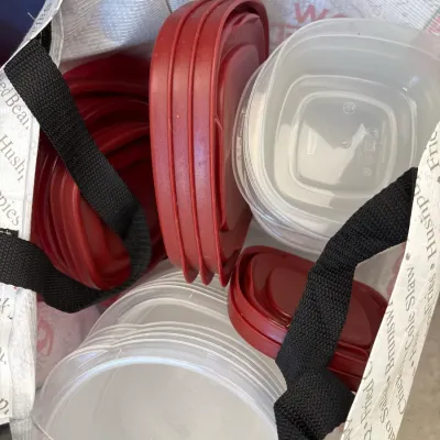 Reusable shopping bag filled with assorted plastic food storage containers and red lids.
