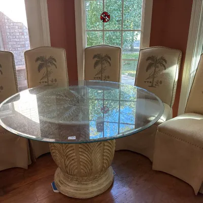 Tropical style round glass-top dining table with a carved palm-motif pedestal base, surrounded by five matching upholstered dining chairs...