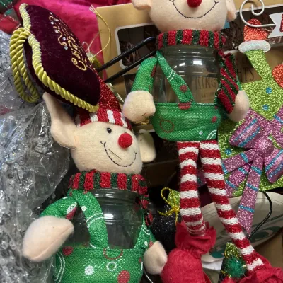 Pair of plush Christmas elf jar covers featuring striped tights, green overalls, and red pointed hats alongside other holiday decor.