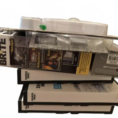 Stack of boxed household items including a Handy Brite cordless work light, Sharper Image items, and an Emerson device.