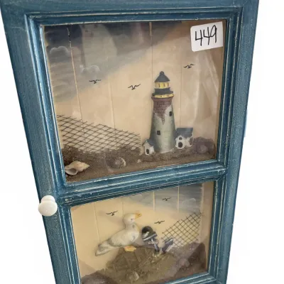 Blue painted wood shadow box display cabinet featuring a nautical theme with a lighthouse, seagull, anchors, and seashells behind glass.