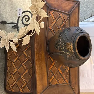 Decorative wooden wall sconce shelf with a woven inlay and urn accent, accompanied by a white wrought-iron floral mirror frame.
