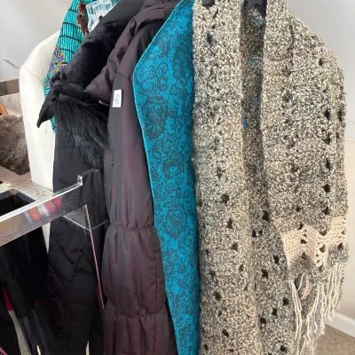 Selection of women's outerwear and knitwear hanging on a metal clothing rack, including a chunky crocheted shawl with fringe and...