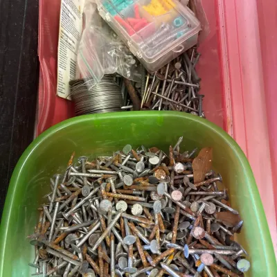 Assorted hardware including loose roofing nails in a green plastic bowl alongside screws, drywall anchors, and wire in a pink storage bin.