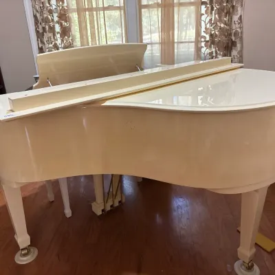 Off-white or cream-colored baby grand piano featuring brass pedals, caster cups, and a high-gloss finish.