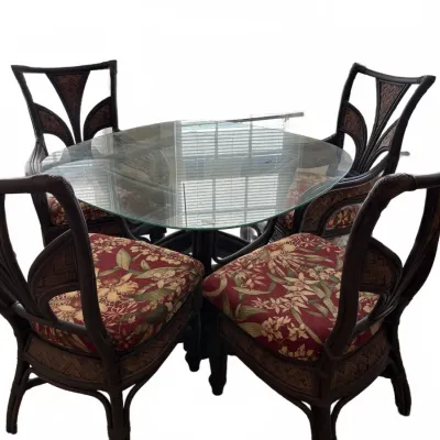 Tropical-style glass-top dining table with four dark rattan armchairs, featuring woven back inserts and floral-patterned seat cushions.