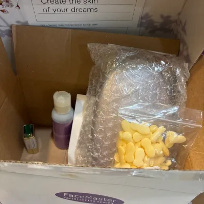 Open cardboard box containing a FaceMaster facial toning system and various beauty supplies, including yellow foam applicator tips and...