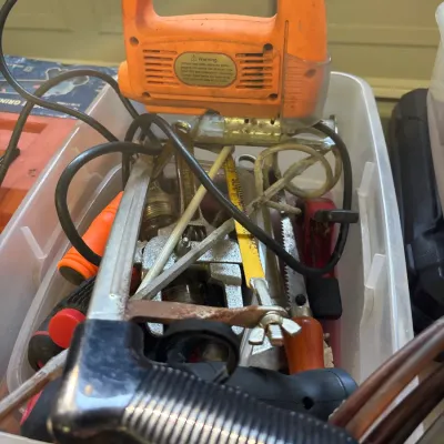 Plastic storage bin filled with mixed hand and power tools, featuring a vintage orange corded jigsaw and a mini hacksaw.