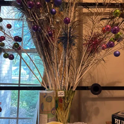 Tall floral and peacock feather arrangement decorated with colorful miniature ornaments and glitter accents in a green glass vase.