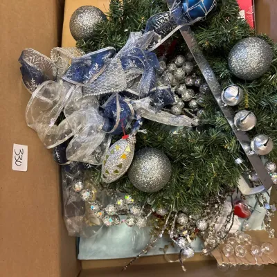 Cardboard box packed with silver and blue Christmas decor, including an evergreen wreath, large glitter ornaments, decorative ribbons,...
