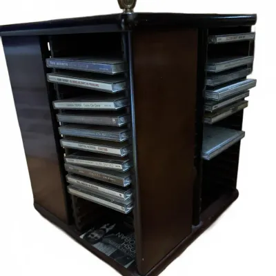 Rotating dark wood CD storage rack filled with various music compact discs, featuring a decorative brass finial on top.