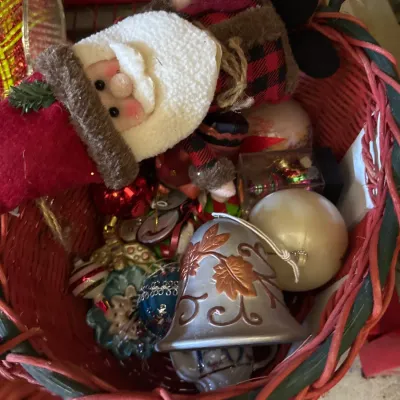 Red and green woven wicker basket filled with mixed holiday decorations, featuring a plush Santa Claus figure, a metallic bell ornament,...
