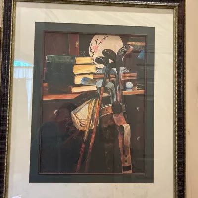 Framed still life print depicting vintage golf clubs, books, and decor, presented in a dark ornate frame with double matting.