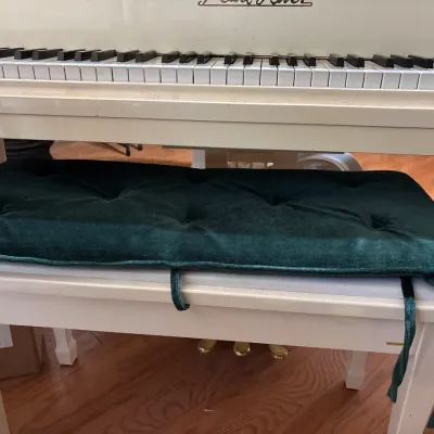 White Pearl River piano bench with a tufted dark green velvet cushion, set beneath the keyboard of a matching white piano.