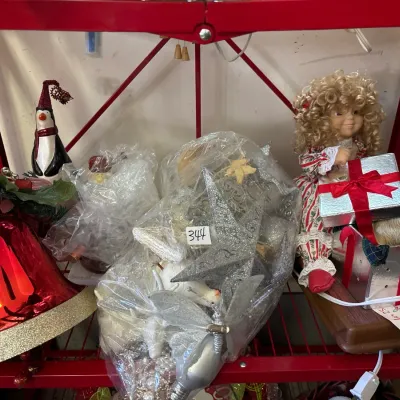 Collection of holiday decorations including large red bell ornaments, a penguin figurine, wrapped glitter stars, and a blonde porcelain...