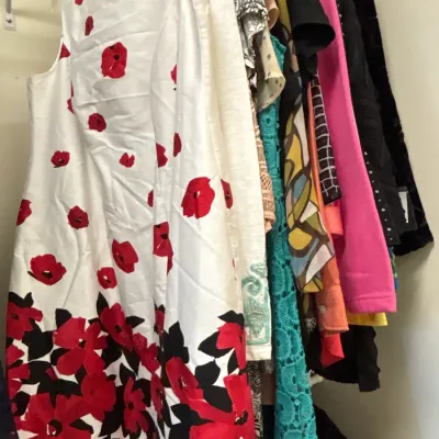 Selection of women's dresses and tops hanging in a closet, featuring a white sleeveless dress with a bold red floral pattern in the...