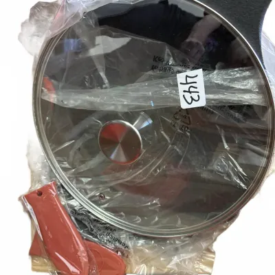 Cast iron skillet set wrapped in original protective plastic, featuring a glass lid with a stainless knob and red silicone handle grips.