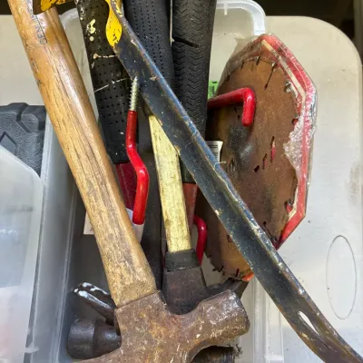 Assorted hand tools in a plastic storage bin, including hammers, a pry bar, storage hooks, and a circular saw blade. The tools show signs...