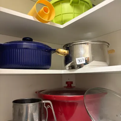 Assorted modern cookware and kitchen tools arranged on pantry shelves, including a large red roaster, cobalt blue covered dish, stainless...