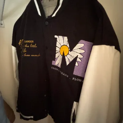 Black and white varsity jacket featuring embroidered details including a large purple 'M' with a daisy graphic, yellow text,...