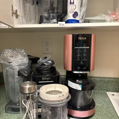 Collection of modern kitchen appliances, including a rose gold Ninja Creami ice cream maker, Ninja blenders with various pitcher...