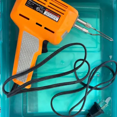Bright orange 180W electric soldering gun kit with attached power cord resting inside a teal plastic storage bin.