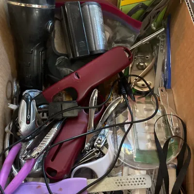 Cardboard box filled with assorted household items and tools, including a flashlight, lighters, pliers, nail files, and hardware...